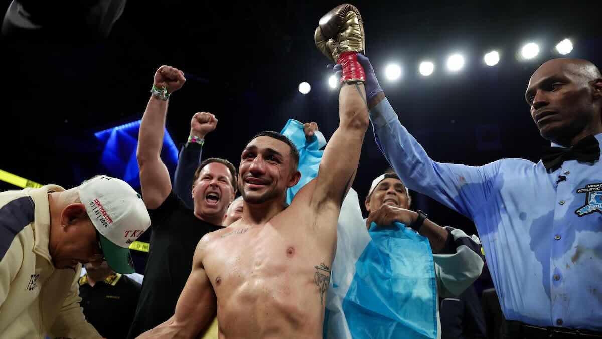Teofimo Lopez 3 Boxer with arm raised in victory as teammates celebrate in the ring at a nighttime bout