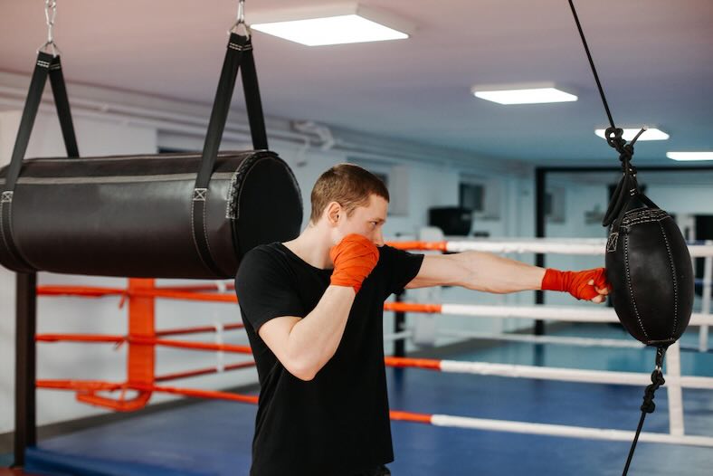 boxeador entrenando Man in a black t-shirt punches a hanging heavy bag in a boxing gym, wearing orange gloves.