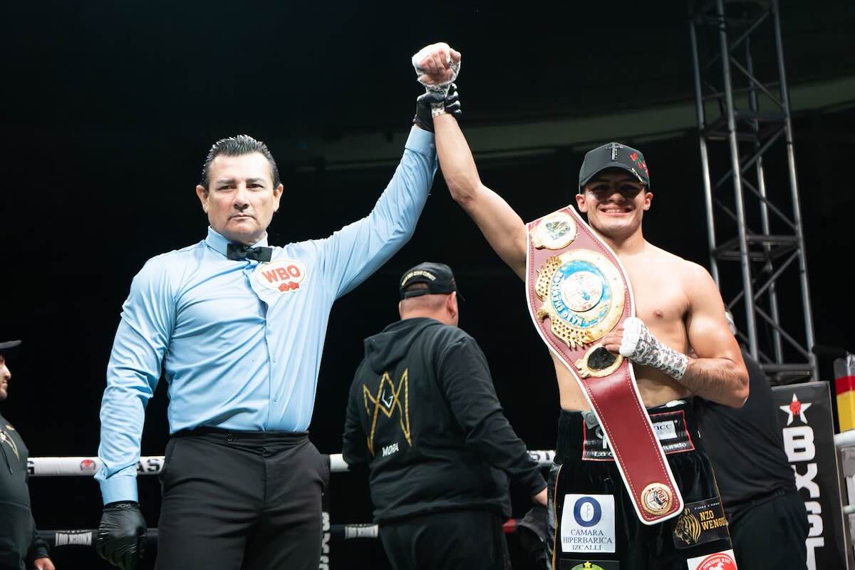 Alberto Mora 1 Referee raises the winner's and his own arms in the boxing ring, the victorious fighter wearing a championship belt and cap smiling.